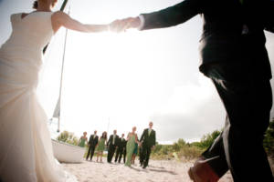 Newly weds holding hands on beach with the wedding party in the background
