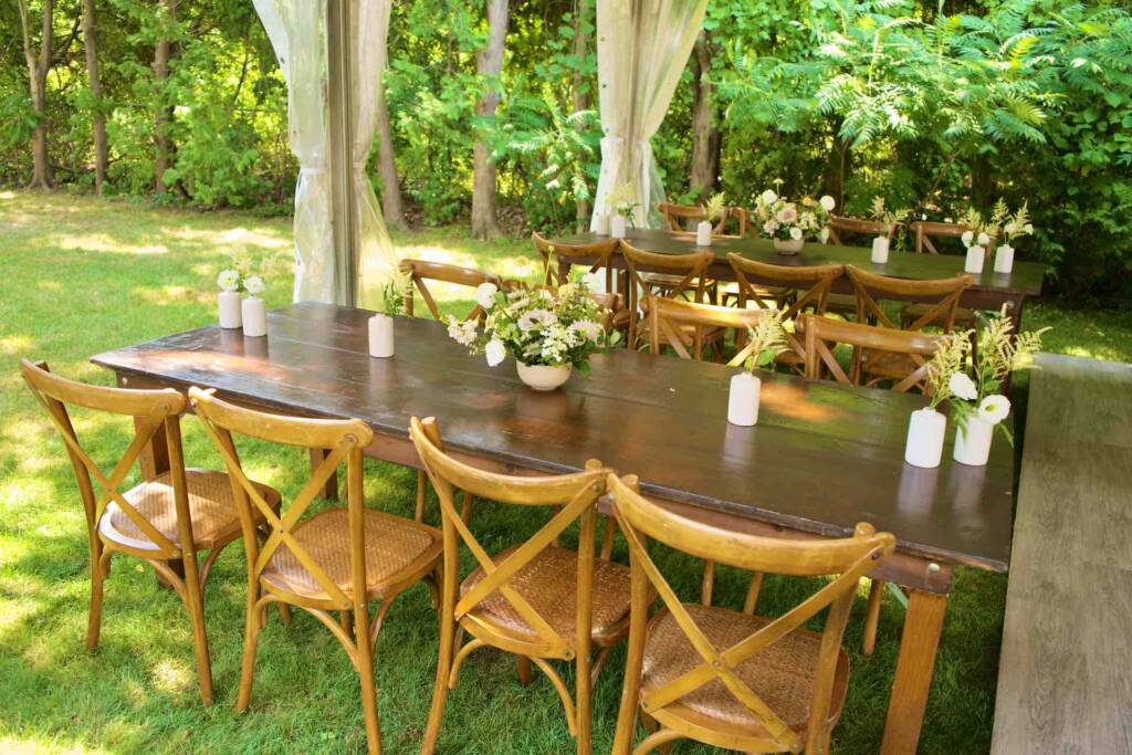 Elegant wood harvest able and chairs set in a grassy backyard under a canopy tent to be used for dinner service for a backyard wedding